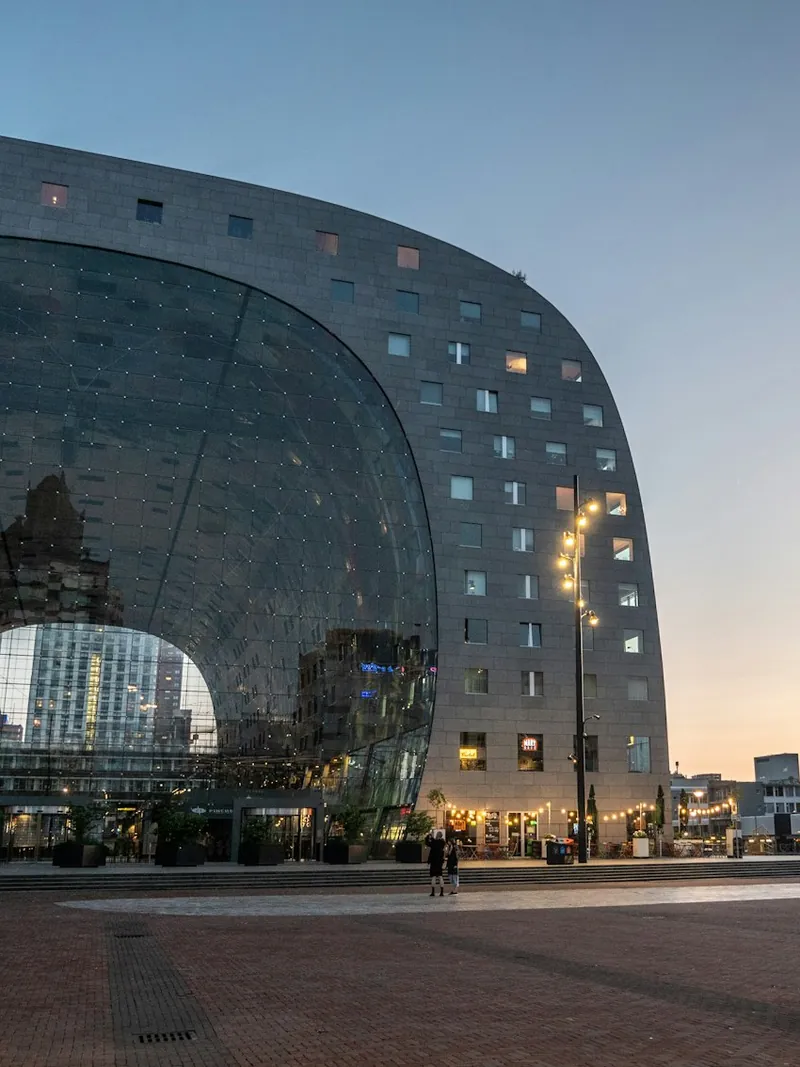 The arched glass front of the Market Hall in Rotterdam at dusk, lit from inside above the empty brick square