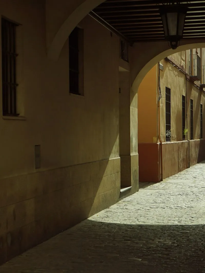 A narrow arched alleyway in Sevilla with ochre walls and a single shaft of sunlight falling across the cobblestones